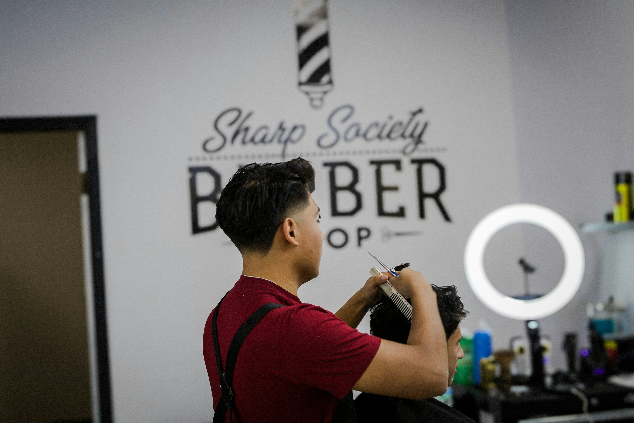 A barber at work in his own shop, cutting a client's hair beneath the shop's painted sign.