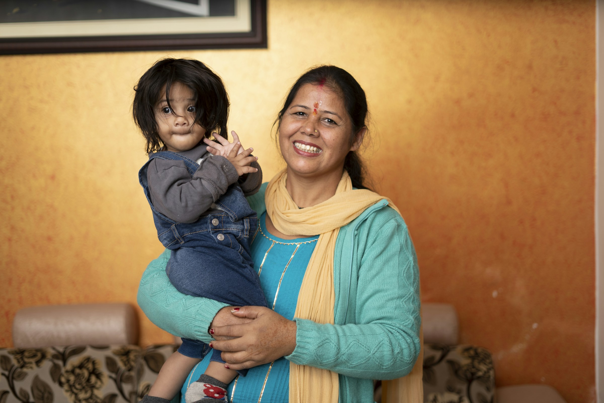 A woman in a teal kurta and yellow dupatta holding her toddler, both looking toward the camera.