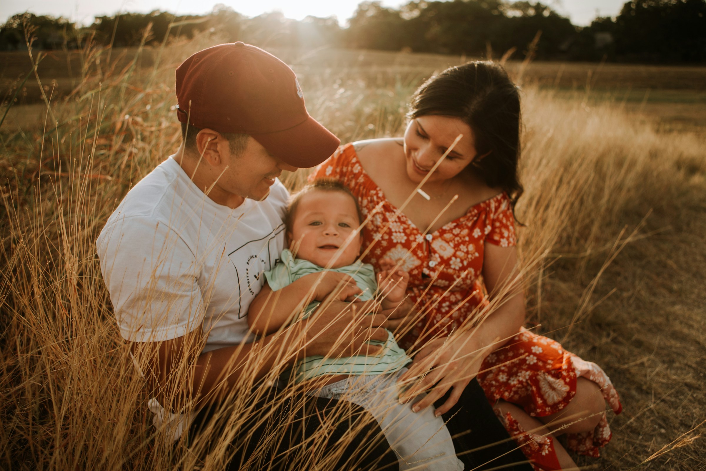 A young family sitting together in tall grass at sunset, the father holding their baby.